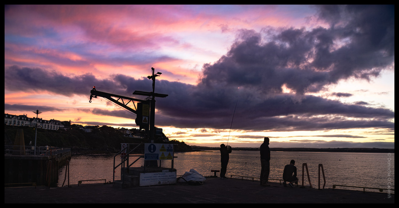 Dramatic sunset scene at Ballycotton harbour showing silhouetted figures fishing from a pier with a harbour crane and colourful pink and purple clouds reflected in the water, County Cork.