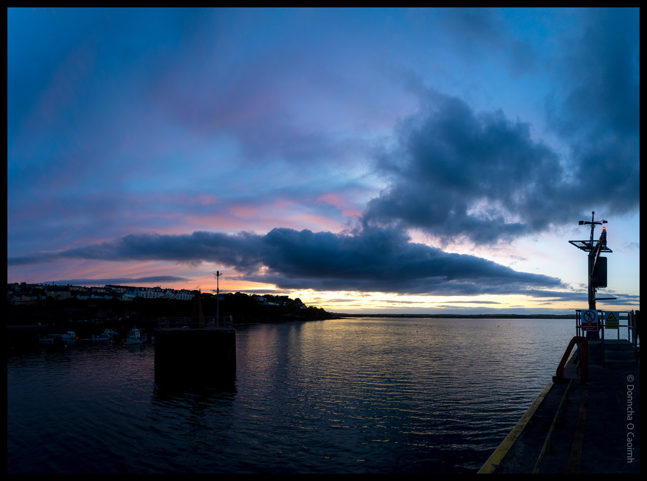 Dramatic twilight scene at Ballycotton harbour, showing dark clouds illuminated by pink light from the setting sun below the horizon, with the village visible across calm water and harbour infrastructure in the foreground.