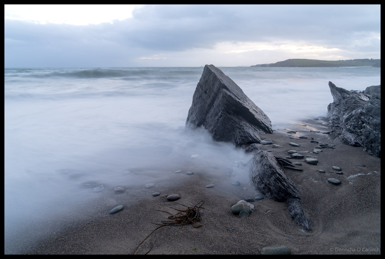 Long exposure photograph of waves washing around large weathered rocks on Owenahincha Beach, creating misty white water effects with scattered stones and seaweed on the sand under an overcast sky, County Cork.