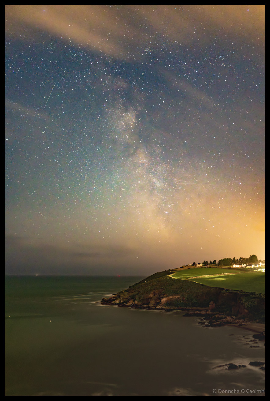 Astrophotography image showing the Milky Way galaxy stretching across a star-filled night sky above the rocky coastline and beach of Church Bay, County Cork, with meteor streaks visible and coastal lights in the distance.