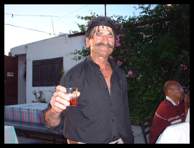 Middle-aged man with distinctive moustache and headband holding a small glass of red drink at an outdoor evening party in a Cretan village, with white buildings and flowering trees in the background.
