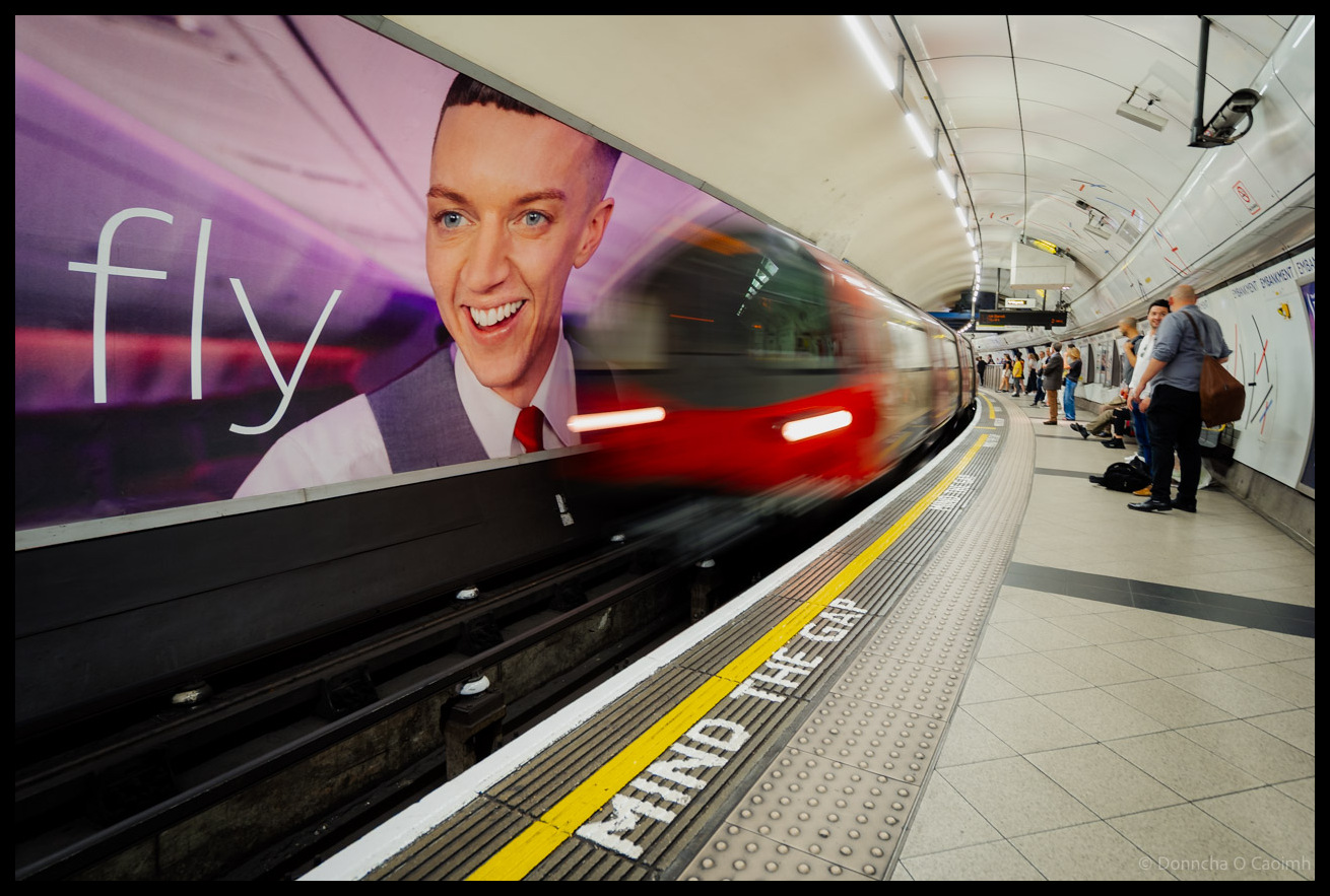 Underground railway platform in London showing motion-blurred tube train with red lights passing large advertisement displaying "fly" text and smiling man in suit, with "MIND THE GAP" warning visible on platform edge and passengers waiting.