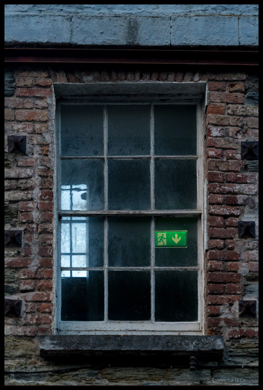 Weathered red brick building facade in Kinsale, Co Cork showing Georgian-style sash window with white painted frame and green emergency exit sign with running figure and downward arrow visible in first floor window