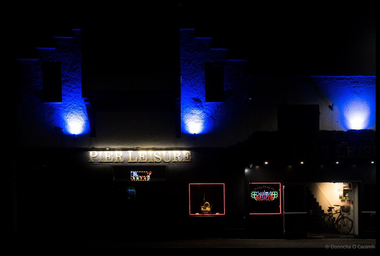 Night-time photograph of Pier Leisure building in Kinsale, Co Cork with dramatic blue uplighting illuminating white chimneys, illuminated "PIER LEISURE" signage, red-framed windows, neon signs, and a bicycle visible in the lit doorway