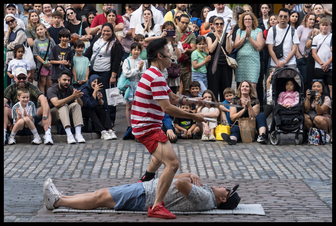 Street performer in red and white striped shirt and red shorts performing acrobatic juggling act with knives while balancing on person lying on mat, surrounded by large crowd of tourists and families watching in Covent Garden, London.