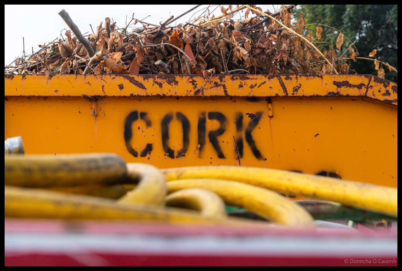Close-up of orange skip or container with "CORK" stencilled in black letters, filled with dried autumn leaves and garden waste, with yellow hoses visible in foreground at Ballincollig Regional Park allotments.