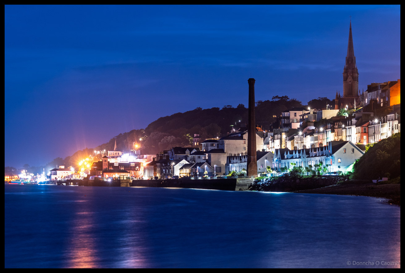 Night-time view of Cobh, Ireland, with the calm waters of Cork Harbour in the foreground, brightly lit houses lining the waterfront, an old red-brick chimney stack standing tall, and St Colman’s Cathedral rising with its illuminated spire against a deep blue sky.