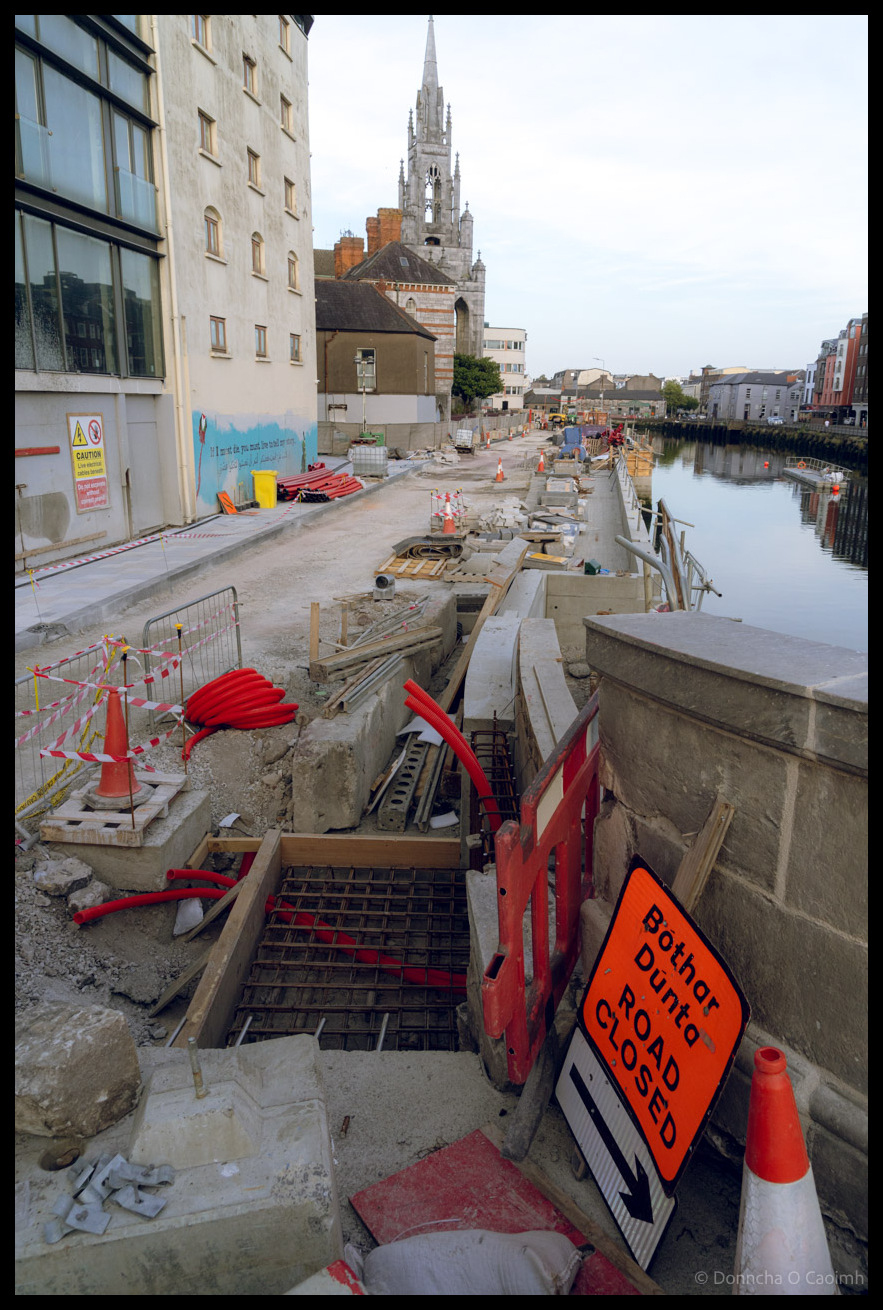 Major roadworks on Father Mathew Quay showing excavated trenches with steel reinforcement, construction equipment, and "Bóthar Dúnta / Road Closed" bilingual warning signs, with Holy Trinity Roman Catholic Church's spire visible in the background, Cork City.