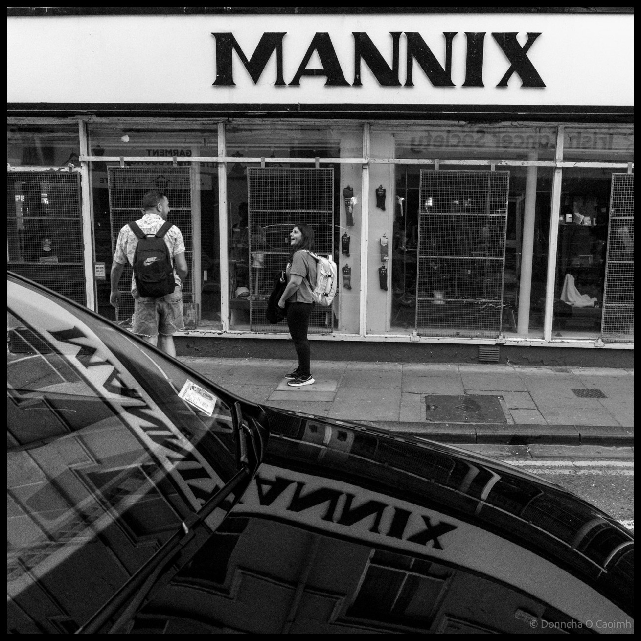 Black and white street photograph showing two people with backpacks walking past Mannix shop on Castle Street Cork, with the shop's name reflected in a curved car windscreen in the foreground.