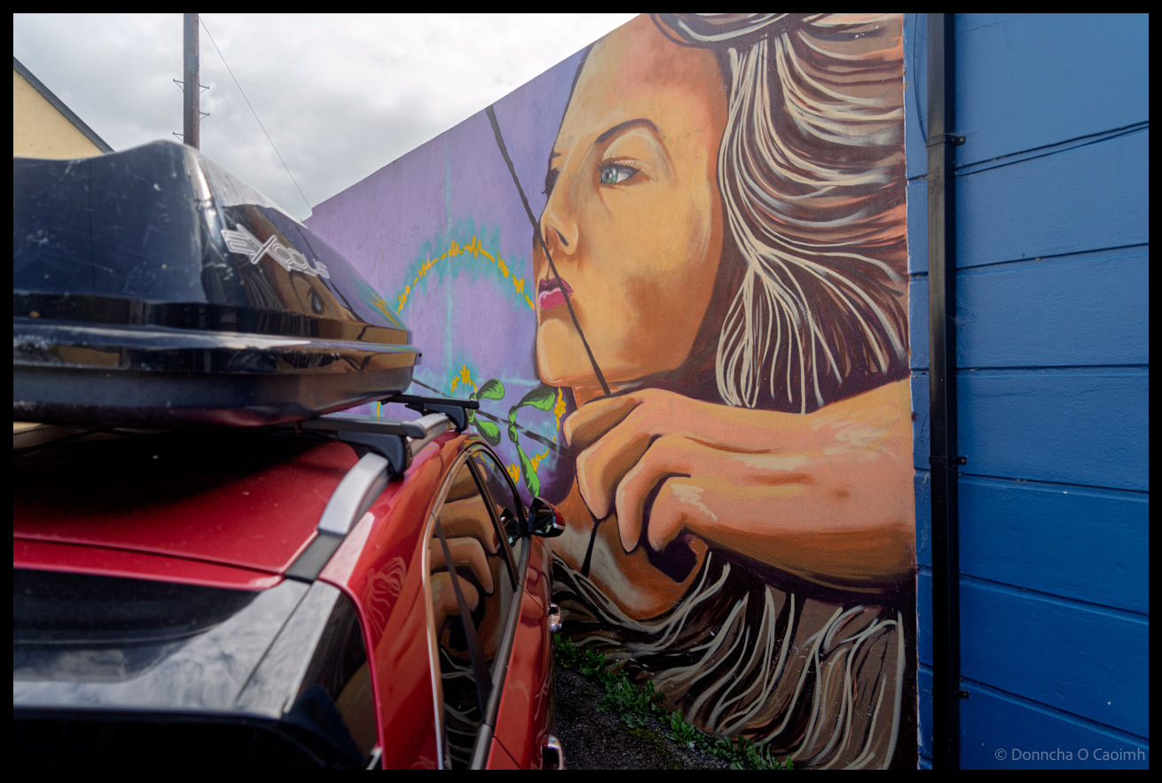 Large-scale street mural on blue wall showing portrait of woman with flowing blonde hair and blue eyes with a bow and arrow made with a sycamore-seed tip. A red car parked in the foreground on Dykegate Street, Dingle, Co Kerry