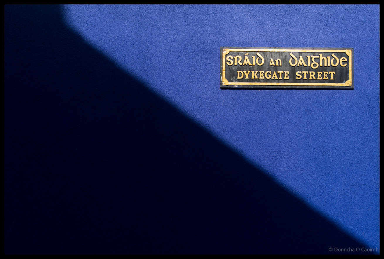 Bilingual street sign reading "SRÁID AN DAIGHIDE / DYKEGATE STREET" mounted on a vibrant blue textured wall with dramatic shadow cast across the surface in Dingle, County Kerry