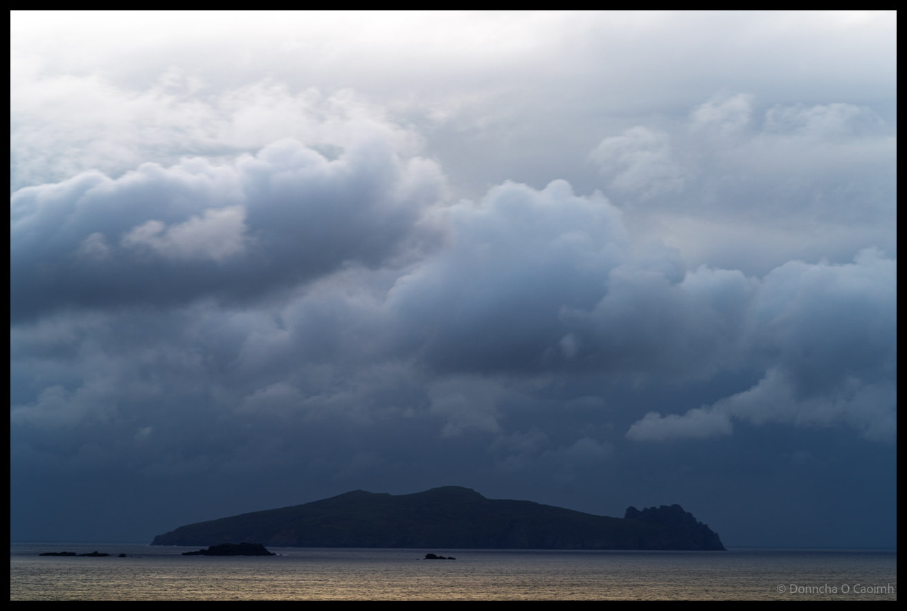 Dramatic storm clouds gathering over the silhouetted form of An Fear Marbh (The Dead Man) rock formation viewed across dark waters from the Dingle Peninsula, County Kerry.