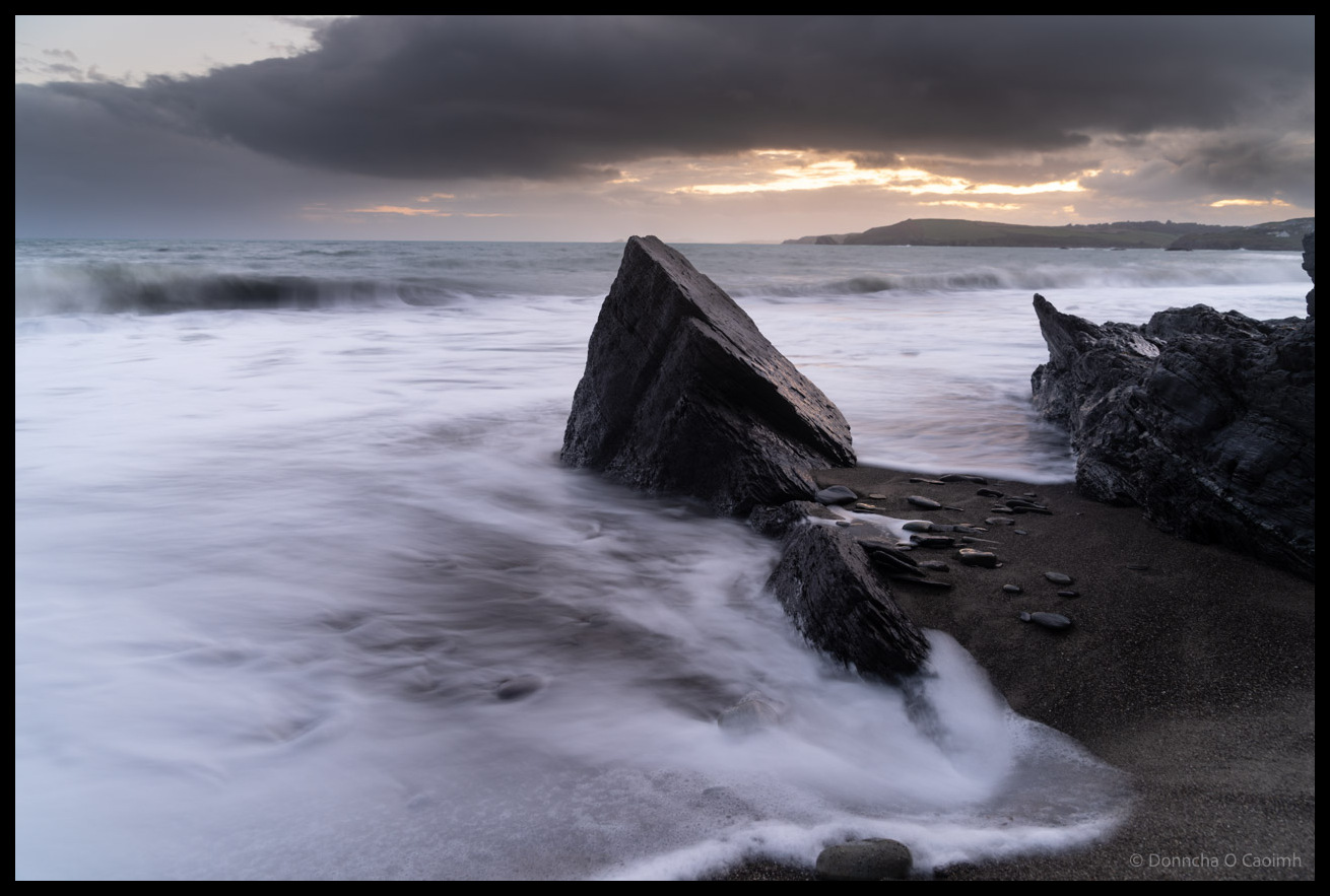Dramatic long exposure seascape at Owenahincha, Co Cork showing weathered rocks and driftwood on dark sand beach with silky motion-blurred waves under stormy sky with dramatic light breaking through heavy clouds over distant headland.