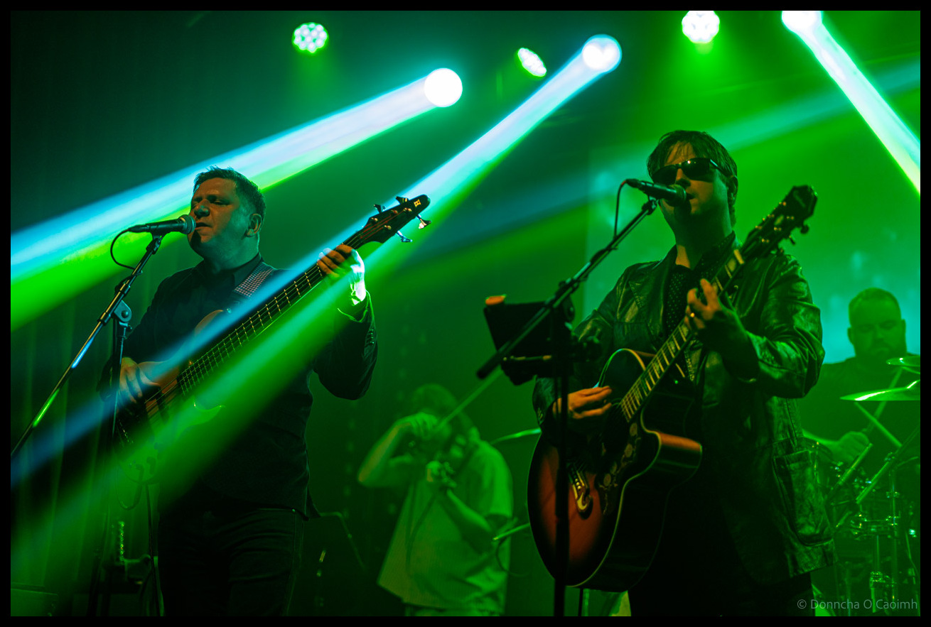 Two musicians from The ELO Experience performing under intense green stage lighting, bassist on left singing into microphone and guitarist on right, both backlit by dramatic concert lighting beams