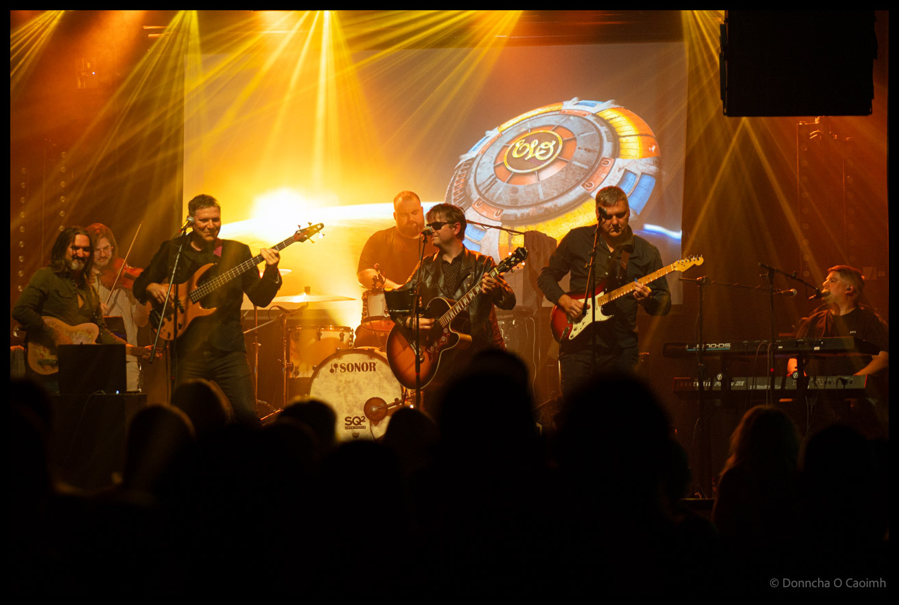 The ELO Experience tribute band performing on stage at Cyprus Avenue Cork with dramatic golden stage lighting, six musicians including bass, guitars, keyboards and drums, with ELO logo backdrop and audience silhouettes in foreground