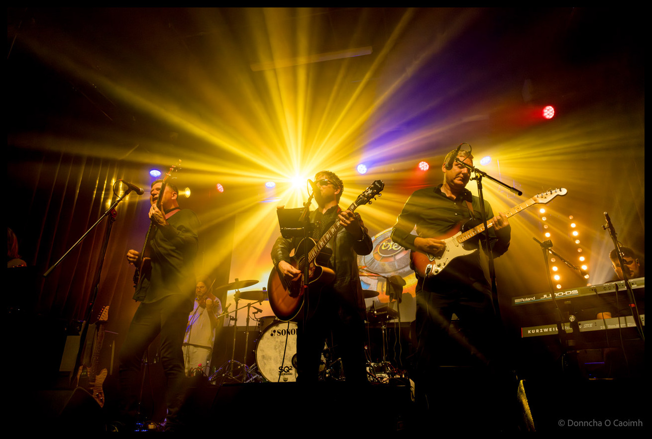 The ELO Experience band on stage with spectacular yellow sunburst lighting effect radiating from centre, three guitarists in foreground with Sonor drum kit visible behind, creating dynamic concert atmosphere