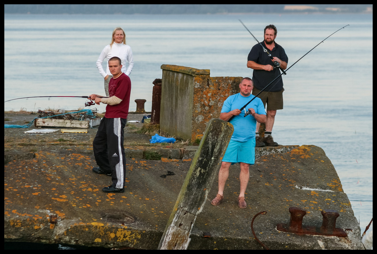 Four people fishing from weathered concrete pier in Cobh, Co Cork with fishing rods, one woman in white top and three men in casual clothing, standing on lichen-covered stone structure with calm harbour waters in background.