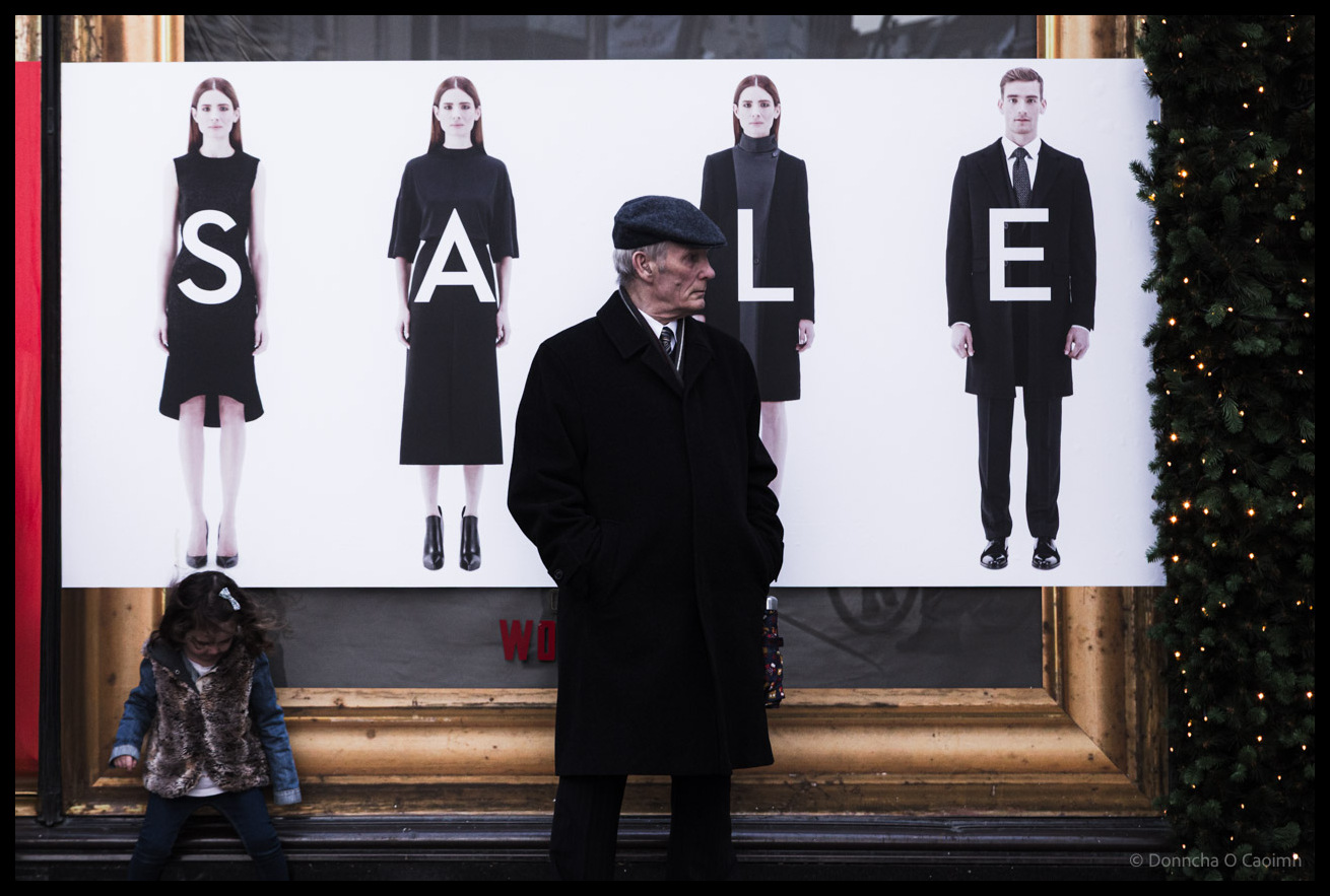 A man wearing a flat cap and long black coat stands in front of a minimalist fashion advertisement displaying models in black clothing spelling out "SALE" at Brown Thomas in Cork, whilst a small girl sits on a ledge below the display with a decorated Christmas tree visible on the right.