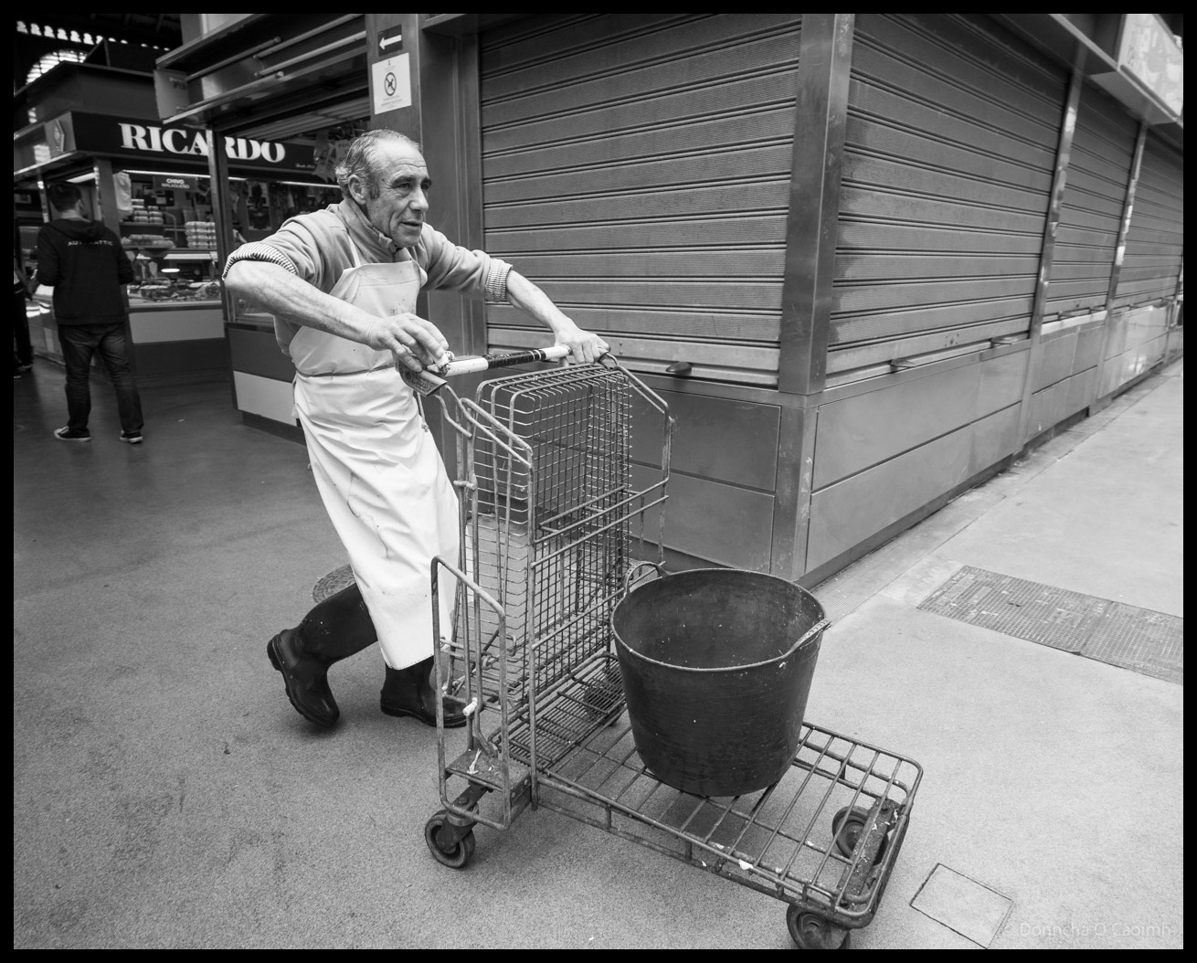 Black and white photograph of a man in white apron pushing a tall wire cage trolley with a large bucket on the lower platform through Atarazanas market in Malaga, Spain, with shuttered market stalls and Ricardo shop visible in the background.