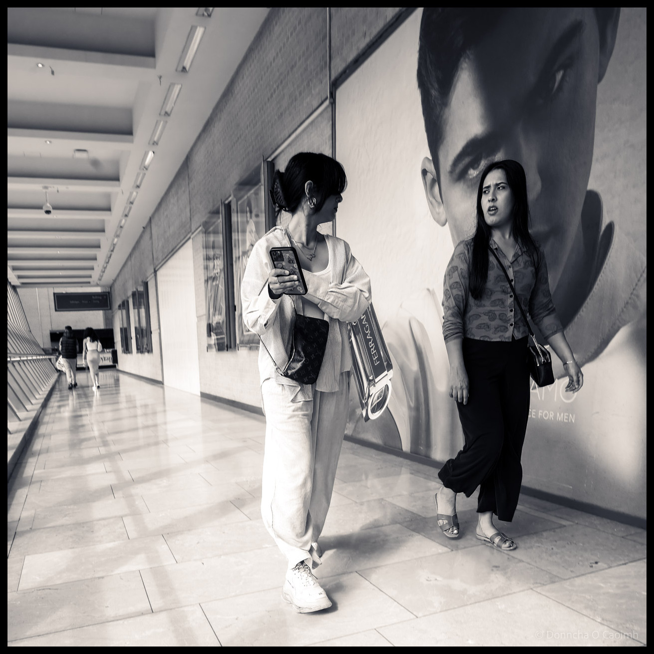Black and white photograph of two women walking through a modern shopping corridor in Birmingham, England, one looking at her phone wearing light-coloured clothing with shopping bags, the other in darker attire standing beside a large fashion advertisement showing a male model's face.