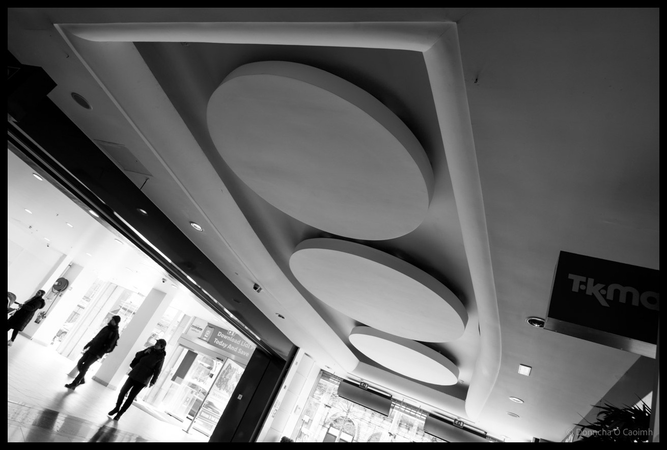 Black and white architectural photograph looking up at distinctive circular recessed ceiling features in The Cornmarket shopping centre in Cork, with silhouettes of shoppers visible through bright storefront windows and a "T.K. Maxx" sign visible on the right.