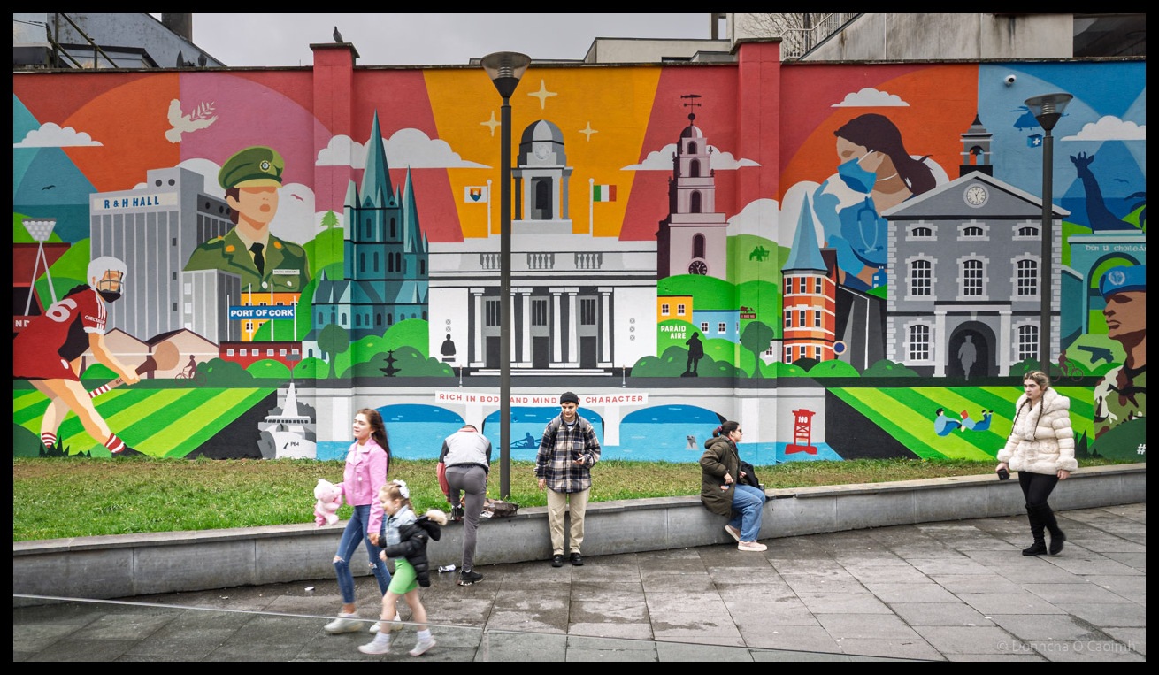 A vibrant large-scale mural just off Paul Street in Cork depicting famous Cork landmarks including City Hall, Shandon bells, Saint Fin Barre's Cathedral, and various Cork icons in a colourful illustrated style, with families and pedestrians sitting and walking along a low wall in front of the artwork.