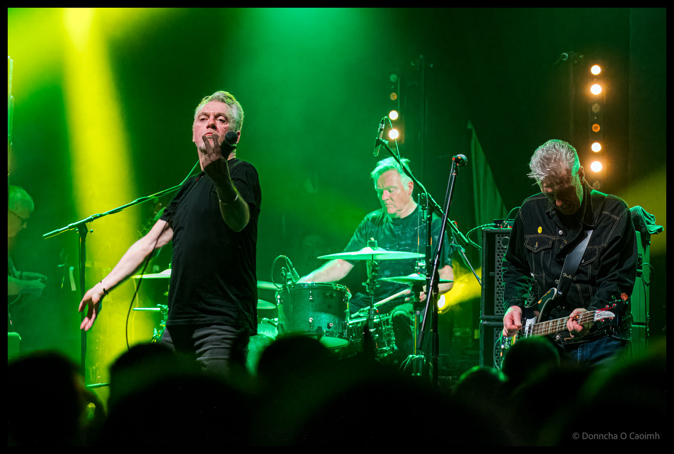 The Undertones performing on stage at Cyprus Avenue in December 2024 under dramatic green and yellow stage lighting, with lead singer at microphone on the left, drummer in the centre background, and bassist on the right, all wearing black, with audience silhouettes visible in the foreground.