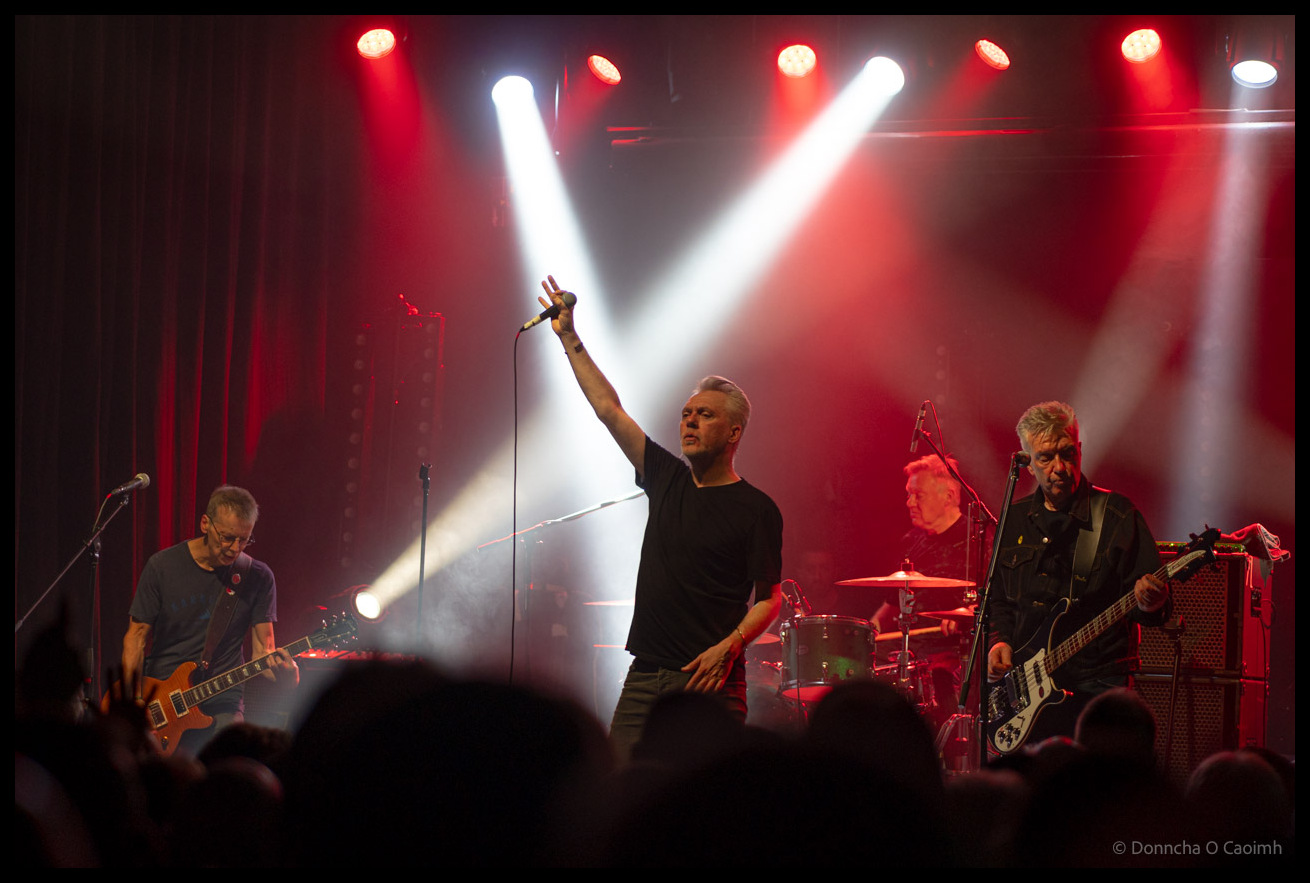 The Undertones performing on stage at Cyprus Avenue with dramatic red and white stage lighting, the lead singer centre stage with arm raised holding a microphone, flanked by a guitarist on the left and bassist on the right, with drummer visible in the background and audience silhouettes in the foreground.