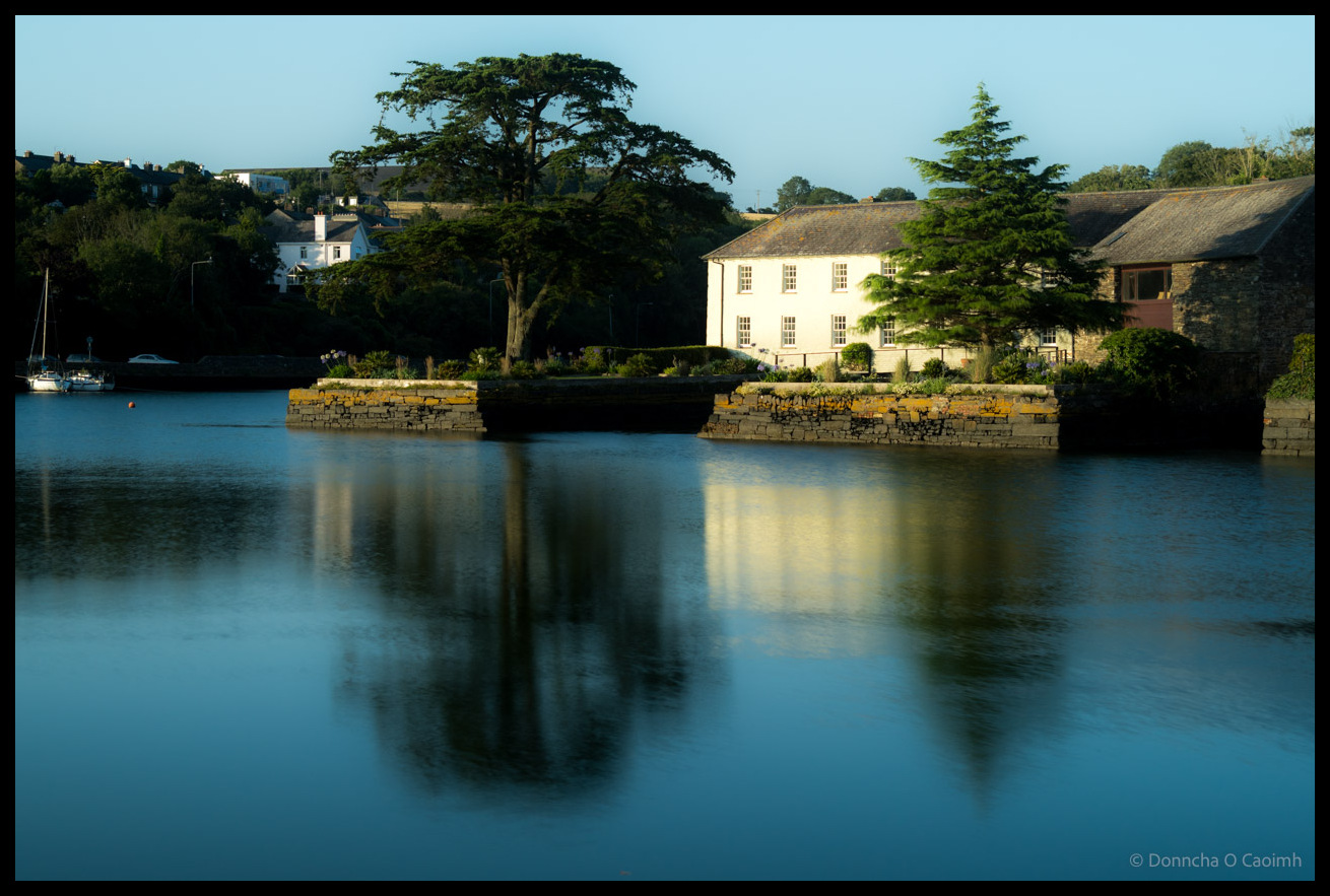 A white Georgian-style house with symmetrical windows sits beside calm blue water in Kinsale, County Cork, with a large mature tree dominating the foreground, stone walls along the waterfront, sailboats moored nearby, and rolling hills with houses visible in the background, all perfectly reflected in the still water.