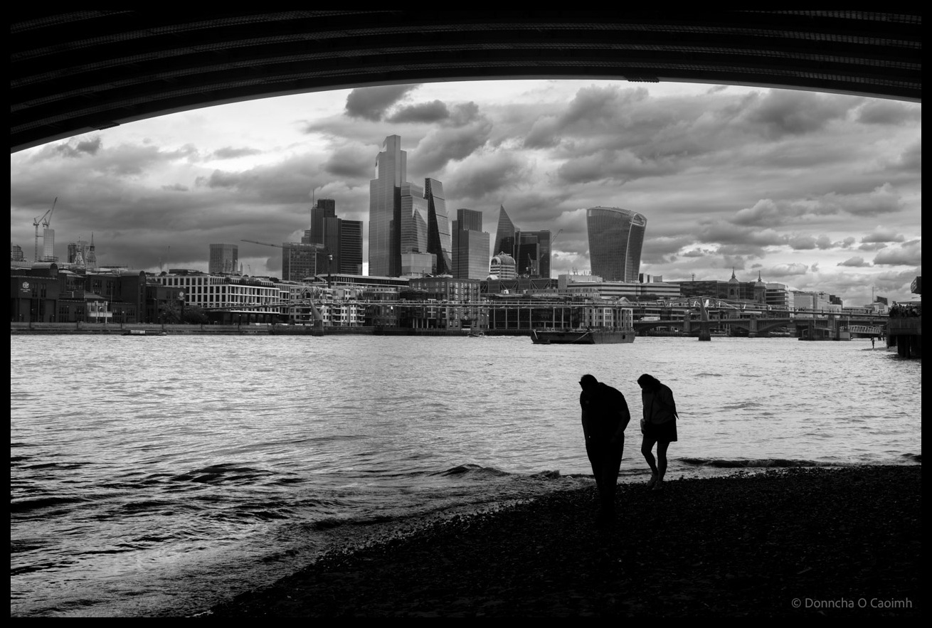 Black and white photograph framed by curved bridge arch of Blackfriars Railway Bridge showing silhouetted couple standing at Thames shoreline with dramatic cloudy sky above London's modern skyline, including the Leadenhall Building, Cheesegrater, and other City skyscrapers across the river.
