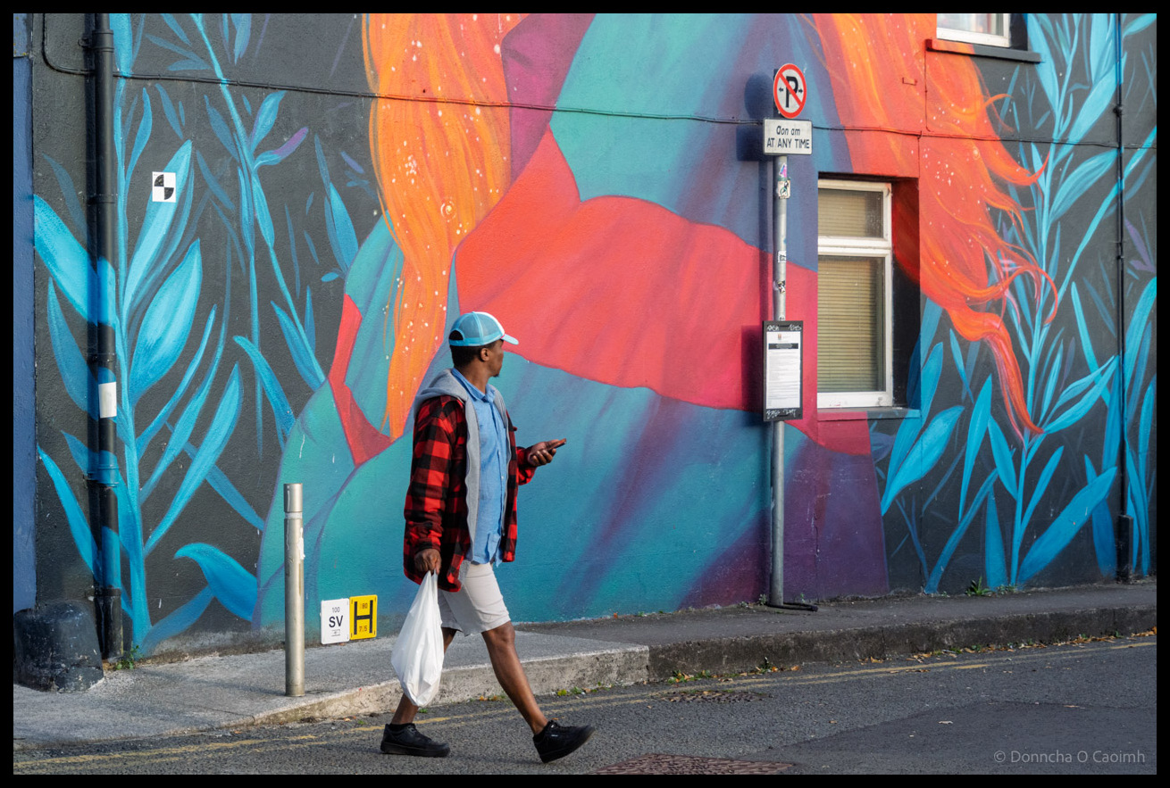 A man wearing a baseball cap, denim gilet, red and black checked shirt, white shorts and carrying a white plastic bag and mobile phone walks past a vibrant street mural featuring green, red and orange shapes and blue foliage on Sullivan's Quay in Cork, with a no parking sign visible on a lamppost.