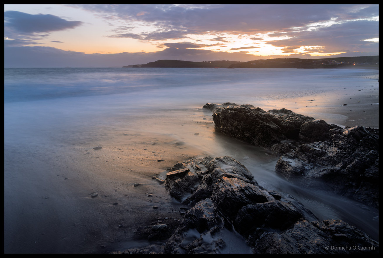 Long exposure photograph of sunset at Owenahincha Beach in County Cork showing smooth, misty water flowing around dark rocky outcrops in the foreground, with golden light breaking through dramatic clouds over silhouetted headlands across the bay.