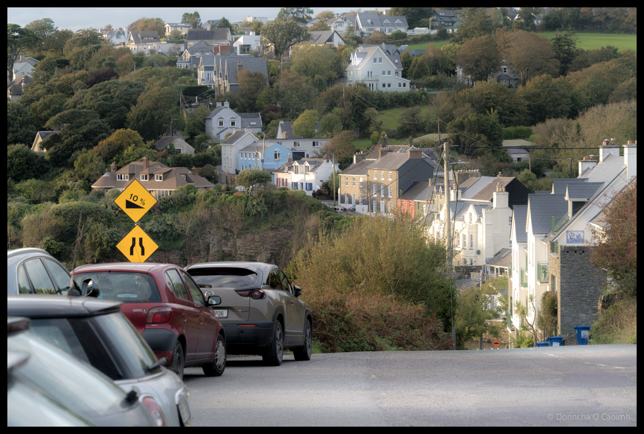 A steep residential street lined with parked cars descends towards a picturesque Irish coastal village with colourful houses nestled among mature trees on a hillside, with two yellow diamond-shaped road signs showing "10%" gradient warning and narrow road ahead symbol in the foreground