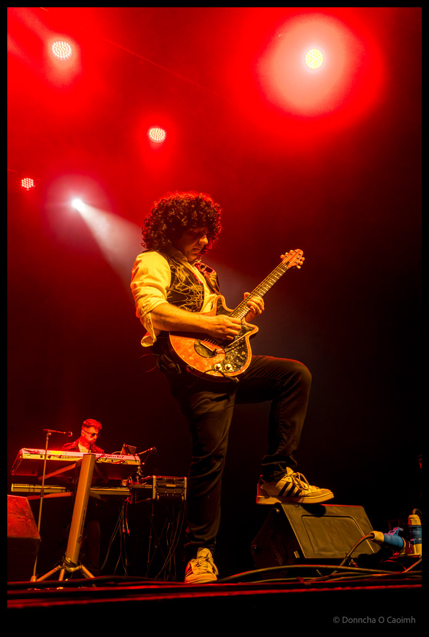 A guitarist with curly dark hair ala Brian May wearing a white shirt made yellow under the red lights and ornate waistcoat playing an electric guitar, with a keyboardist visible in the background at the Cork Opera House during a Qween tribute band performance