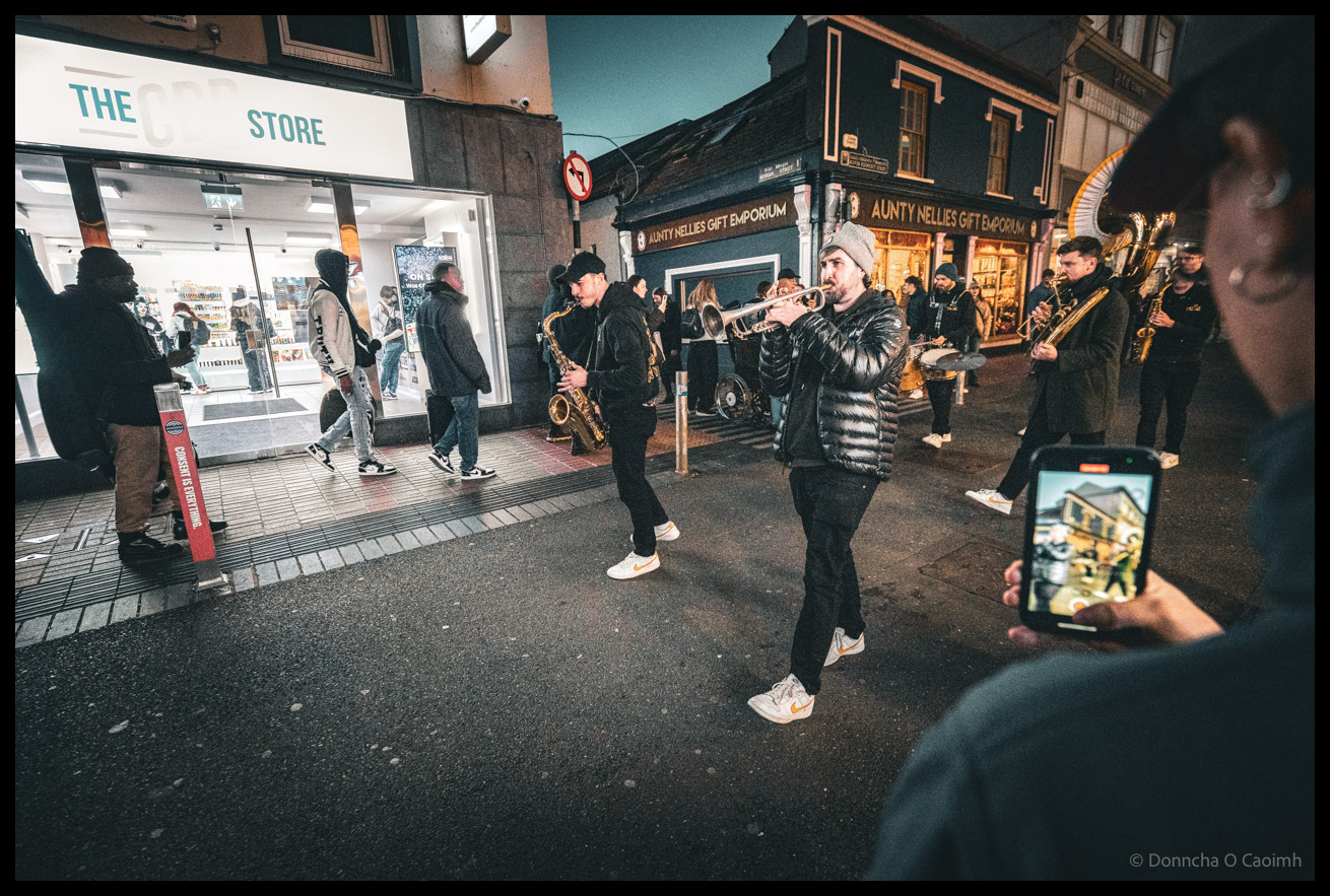 Brass band musicians performing on Patrick Street during Cork Jazz Festival parade at dusk, with trumpeter wearing sunglasses and puffer jacket in centre foreground, surrounded by crowd of spectators many filming on mobile phones, with brightly lit Penneys store and other Patrick Street shops visible in the background under evening sky.