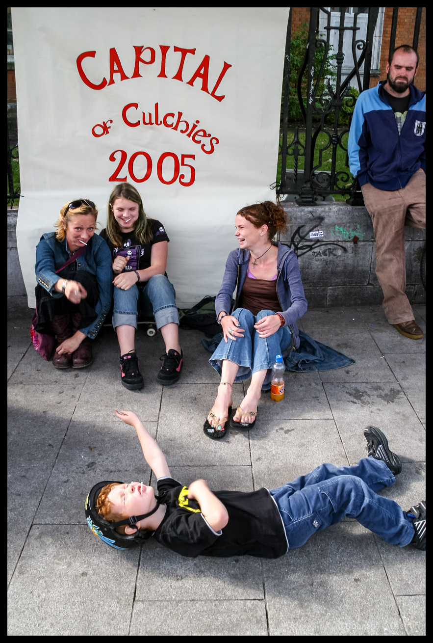 Street scene in Emmett Place, showing three women sitting and chatting in front of a banner reading “CAPITAL of Culchies 2005,” with a man leaning against a railing nearby and a young boy in a helmet lying playfully on the pavement in the foreground. 