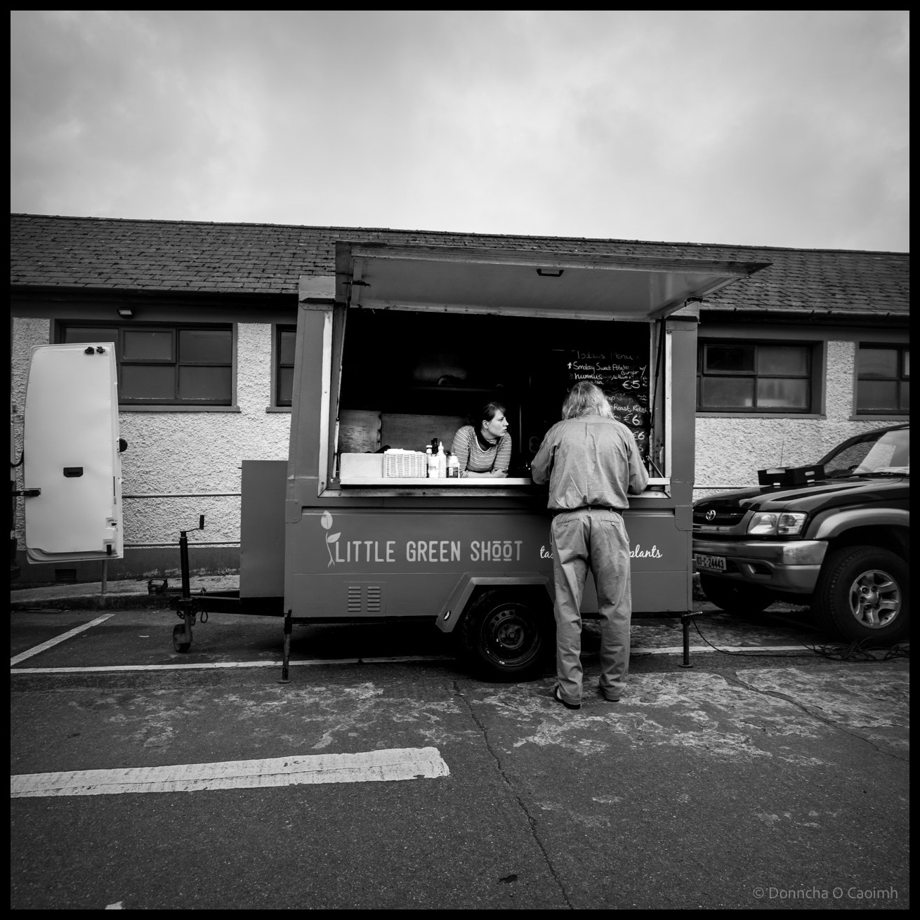 Black and white photograph of a food trailer called "Little Green Shoot" at Skibbereen Market with a customer standing at the service window being served by staff inside, a menu board displaying prices including "€5", surrounded by a parked car on the right and a van door visible on the left, and a single-storey building under overcast sky.