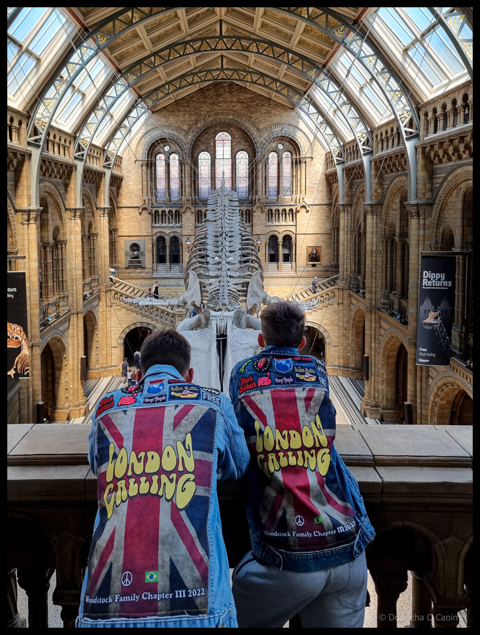 Photograph taken from behind two visitors wearing denim jackets printed with "LONDON CALLING" in yellow text over Union Jack designs and "Woodstock Family Chapter III 2022" patches, standing at a railing overlooking the main hall of the Natural History Museum London with a large skeleton mounted centrally beneath a soaring vaulted glass and timber roof with ornate stone columns and Gothic arches.