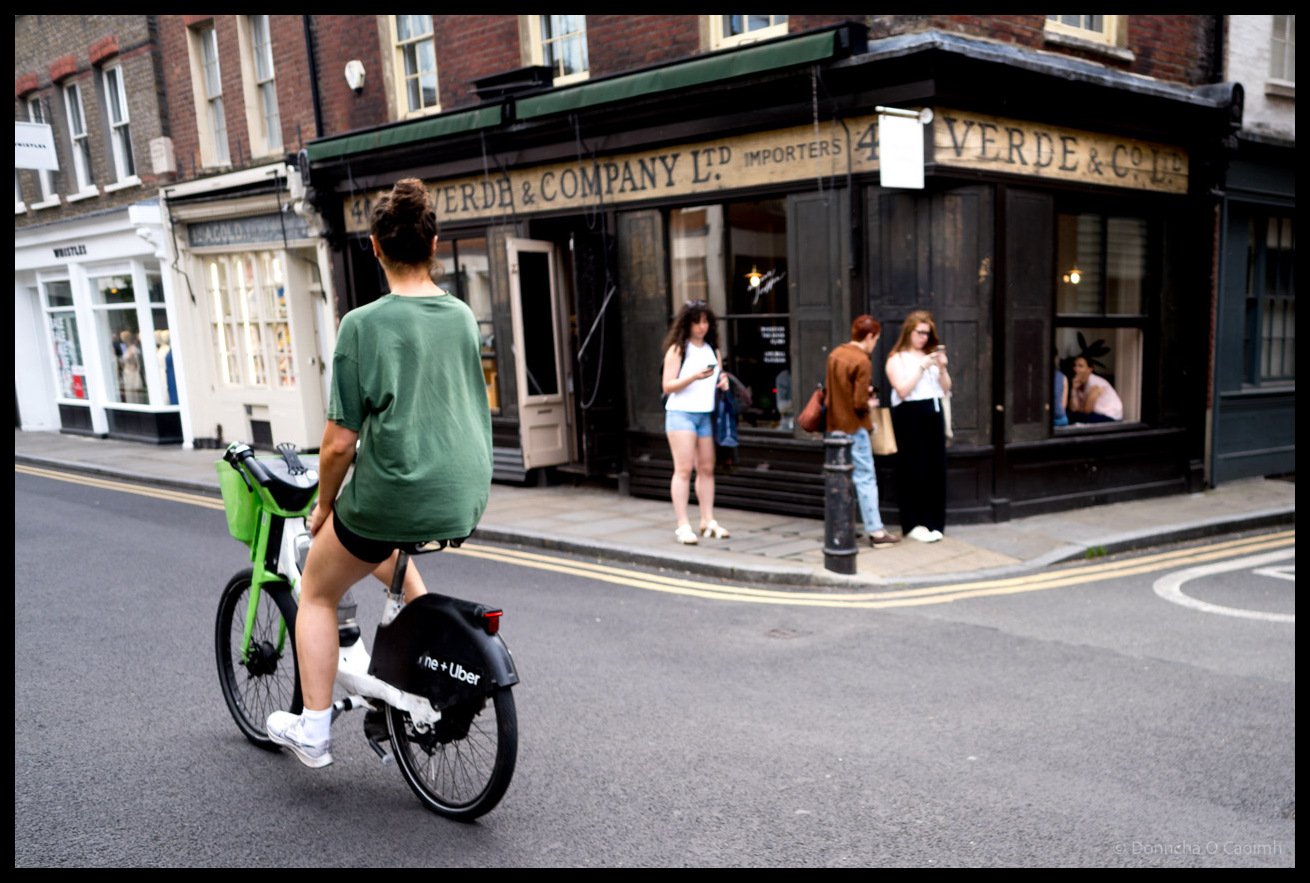 A woman in green t-shirt on a bright-green bike rides past historic black-painted corner shop with gold lettering reading "VERDE & COMPANY LTD IMPORTERS" on a London street, with pedestrians visible near shopfront and period brick buildings in background.