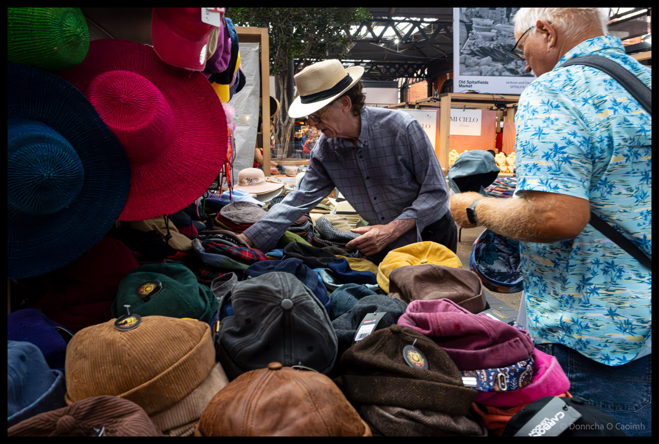 Hat vendor in cream fedora and checked shirt holds out corduroy cap for customer in bright blue tropical print shirt at busy market stall in Spitalfields Market London, with colourful display of caps, berets and fedoras piled on table and vibrant hats hanging above stall.