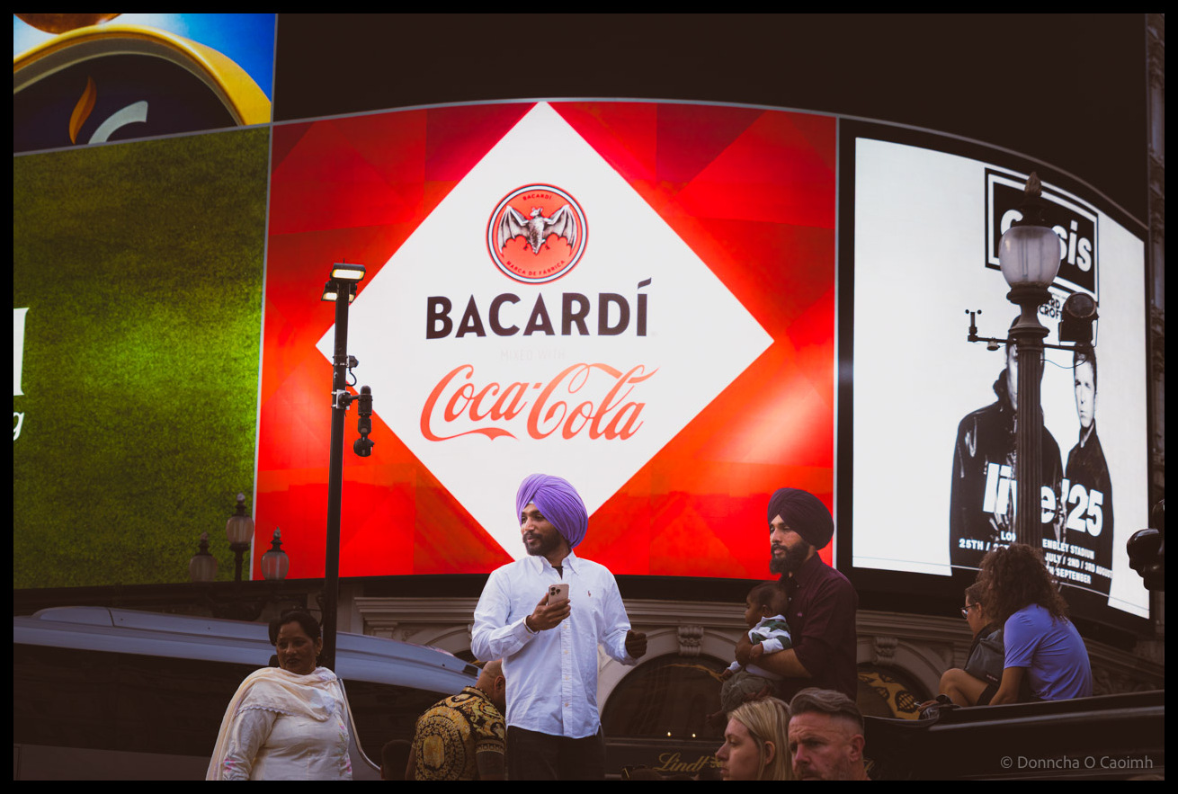 Two Sikh men wearing turbans, one in purple and one in black, standing beneath massive illuminated advertising screens at Piccadilly Circus London showing Bacardi rum and Coca-Cola branding, and an Oasis advert, with diverse crowd of tourists and pedestrians visible below the iconic digital billboards.