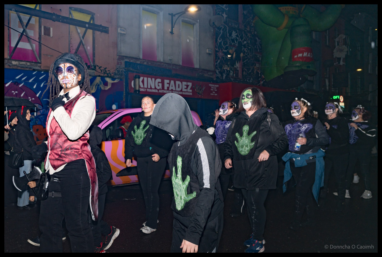 Group of Dragon of Shandon parade participants on Patrick Street in Cork wearing elaborate Day of the Dead face paint and black clothing with green hand designs, including a figure in burgundy vest in foreground and a hooded figure in centre, with colourful murals and King Fadez shop visible in background at night
