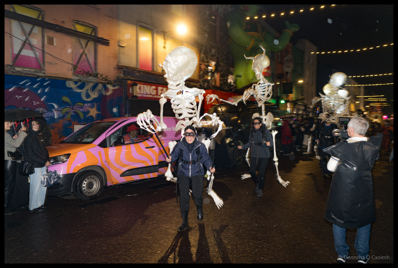 A puppeteer manipulates a large white skeleton puppet on Patrick Street during the Dragon of Shandon parade in Cork, with a pink and orange striped van, costumed parade participants, and spectators visible in the background under street lights at night