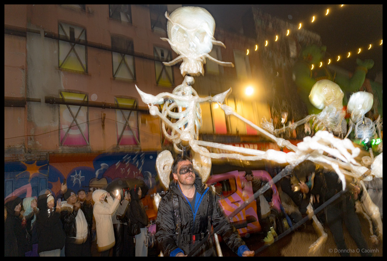 A puppeteer with skull face paint looks up whilst manipulating a towering articulated skeleton puppet overhead during the Dragon of Shandon parade on Patrick Street in Cork, surrounded by crowds of spectators and other costumed participants, with colourful building murals and string lights visible in the background at night
