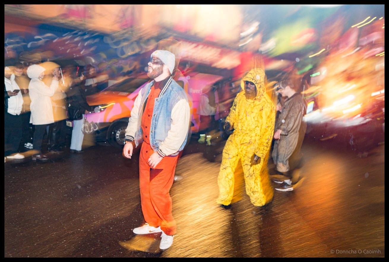 Motion-blurred photograph of Dragon of Shandon parade participants in colourful costumes including orange dungarees, yellow full-body suit, and white beanie on Patrick Street Cork at night, with streaking lights and movement creating dynamic carnival atmosphere.