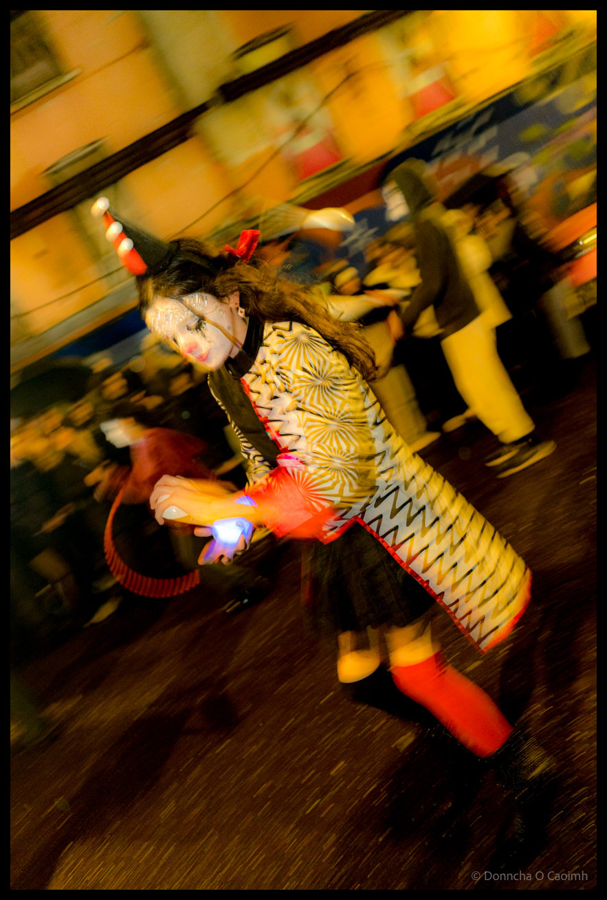 Motion-blurred photograph of young participant in elaborate yellow and red patterned costume with face paint holding illuminated prop during Dragon of Shandon parade on North Main Street, Cork, at night.