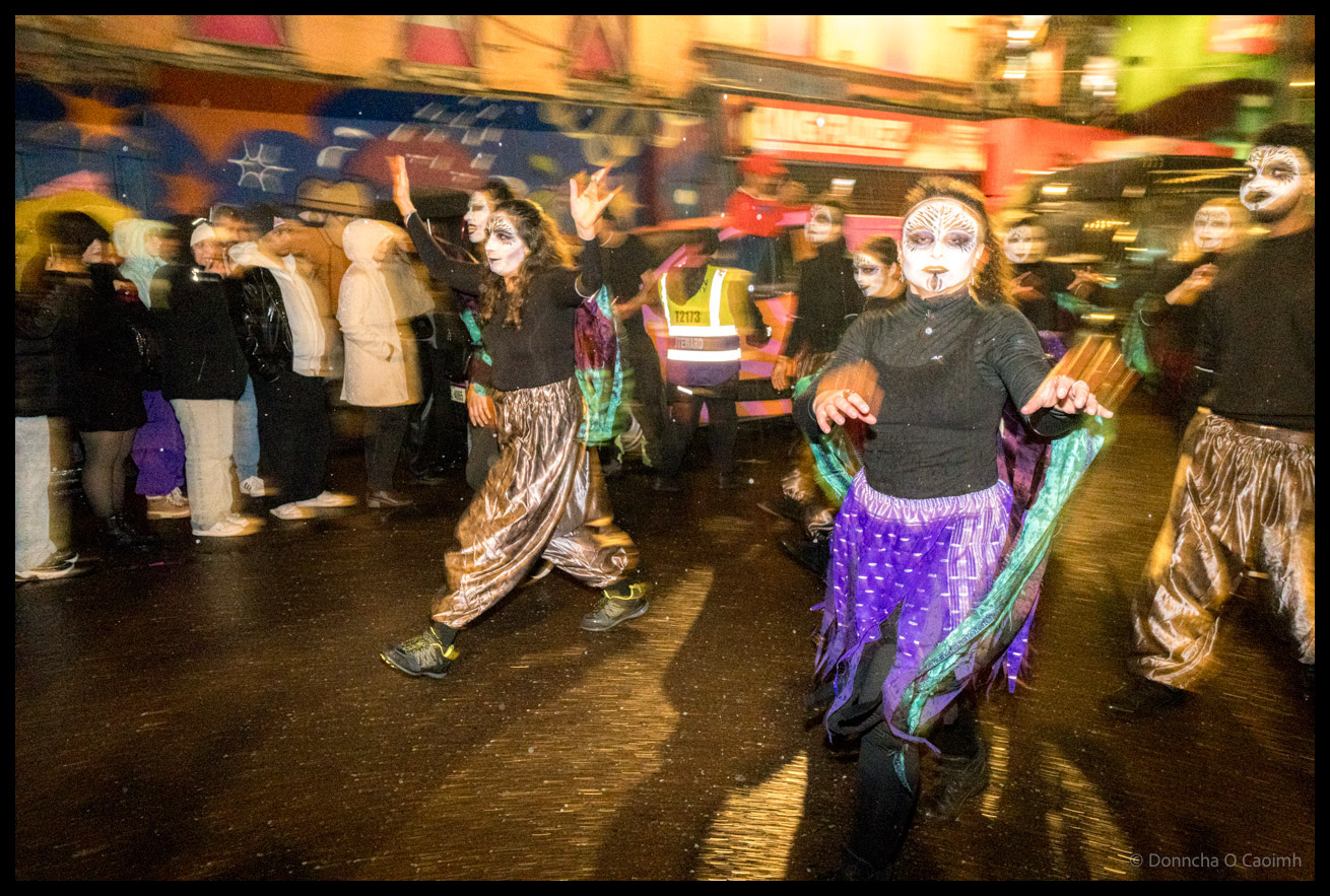 Group of Dragon of Shandon parade dancers in metallic skirts and black clothing with theatrical face paint performing on North Main Street, Cork, at night with colourful murals and parade marshal in hi-vis vest visible in background.