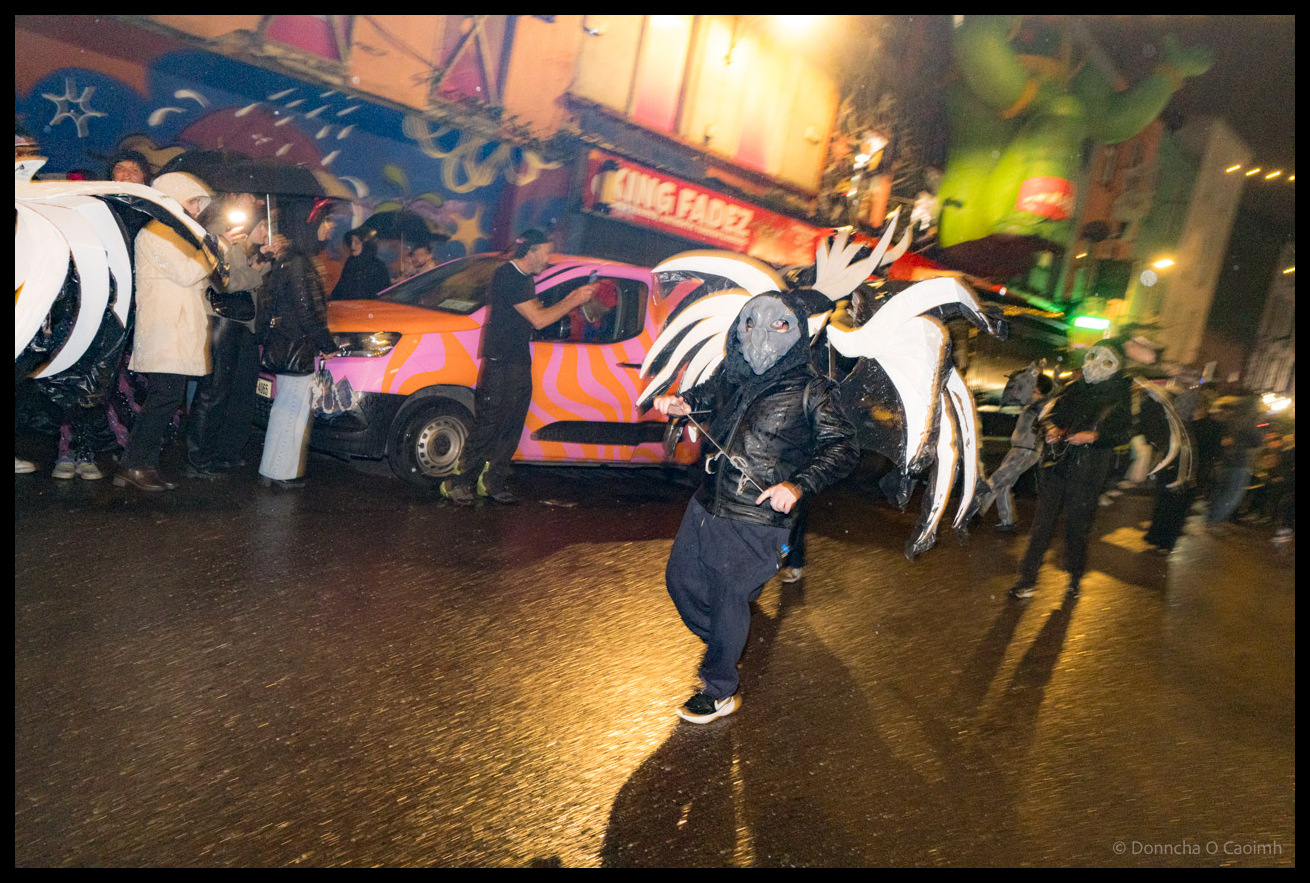 Participant in black feathered bird-like costume with grey mask and large white wings performing dramatic pose during Dragon of Shandon parade on North Main Street, Cork, with pink and orange striped van in background.
