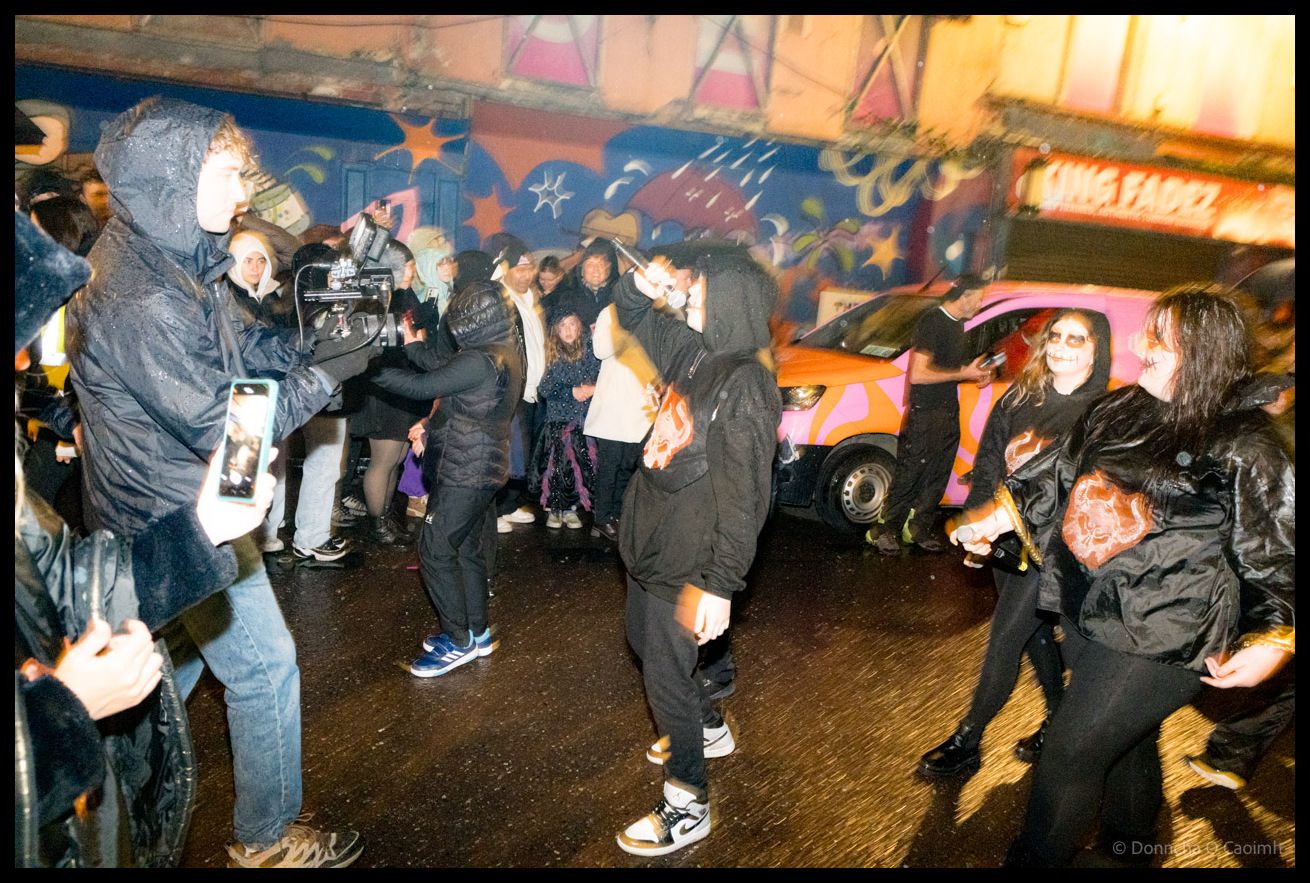 Crowd of Dragon of Shandon parade participants in black clothing and face paint gathered on North Main Street Cork at night with pink and orange striped van and colourful murals visible in background, spectators filming on phones.
