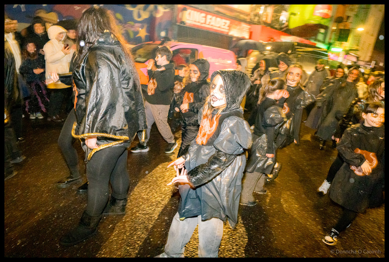 Young participants in elaborate black costumes with skeleton face paint performing choreographed routine during Dragon of Shandon parade on North Main Street Cork, including child in grey outfit in foreground.